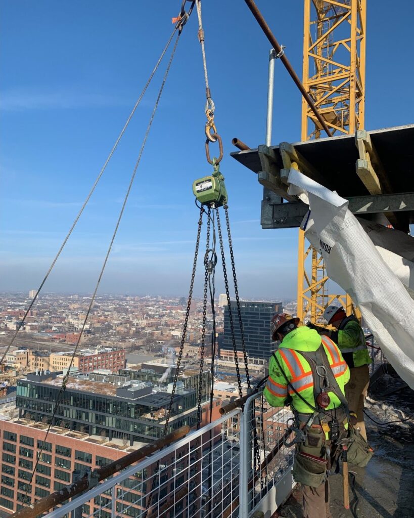 Jobsite photo of a 5 ton electric chain hoist with a construction worker on a platform.